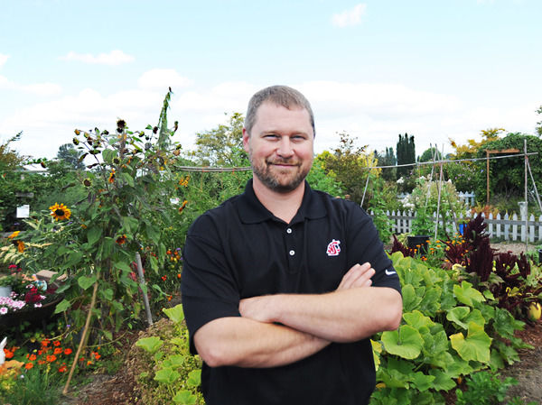 Bearded man stands with arms crossed in front of plants.